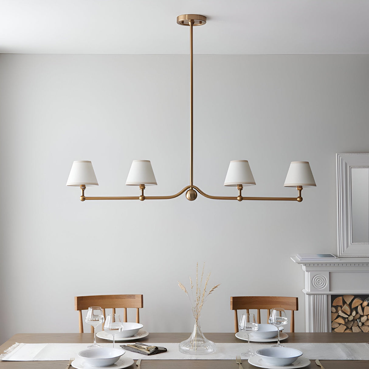 Endon Cliffleigh 4 light linear pendant in brushed aged brass with white tapered fabric shades, hung above a dining table by a fireplace.