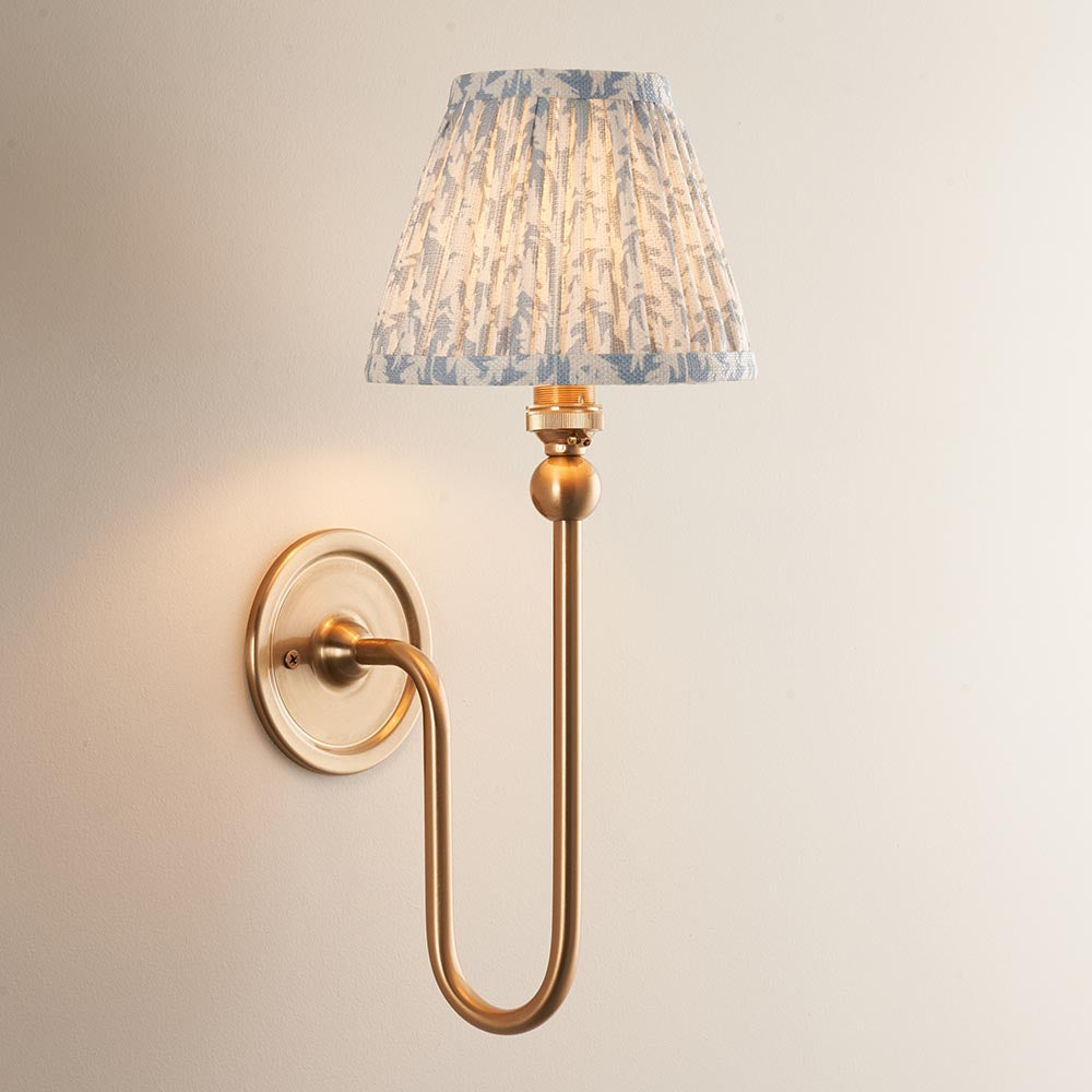 Endon Cliffleigh Leaf wall light in brushed brass with a curved arm and tapered pleated faux linen shade in blue leaf print, on a pale background.