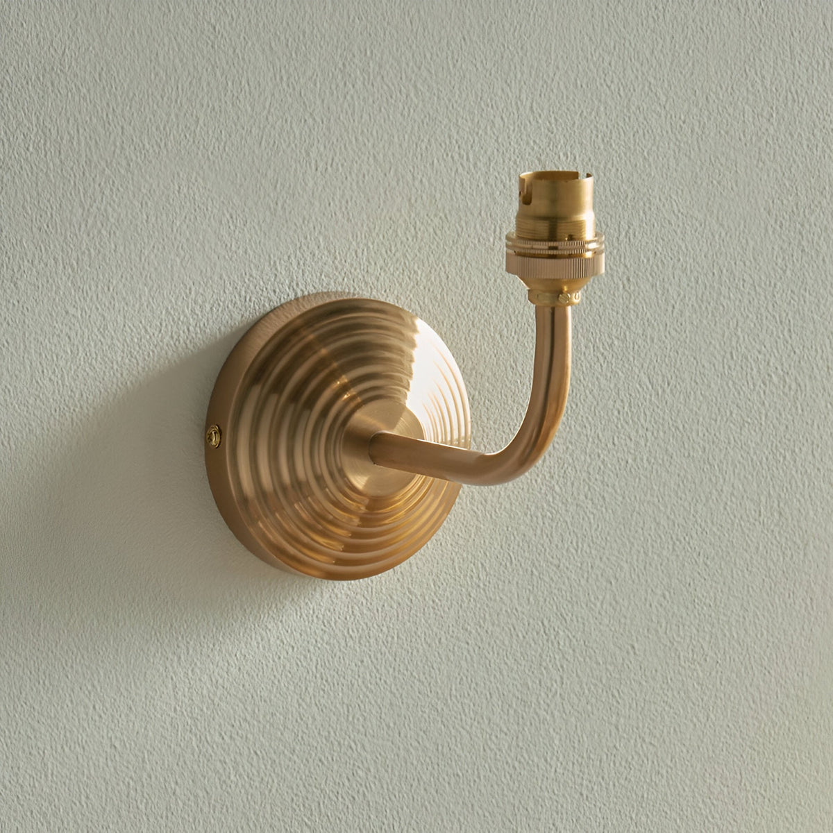 Castgrove Wall Light in brushed aged brass with a stepped circular backplate, curved arm and B22 lampholder, shown against a plain light grey wall.
