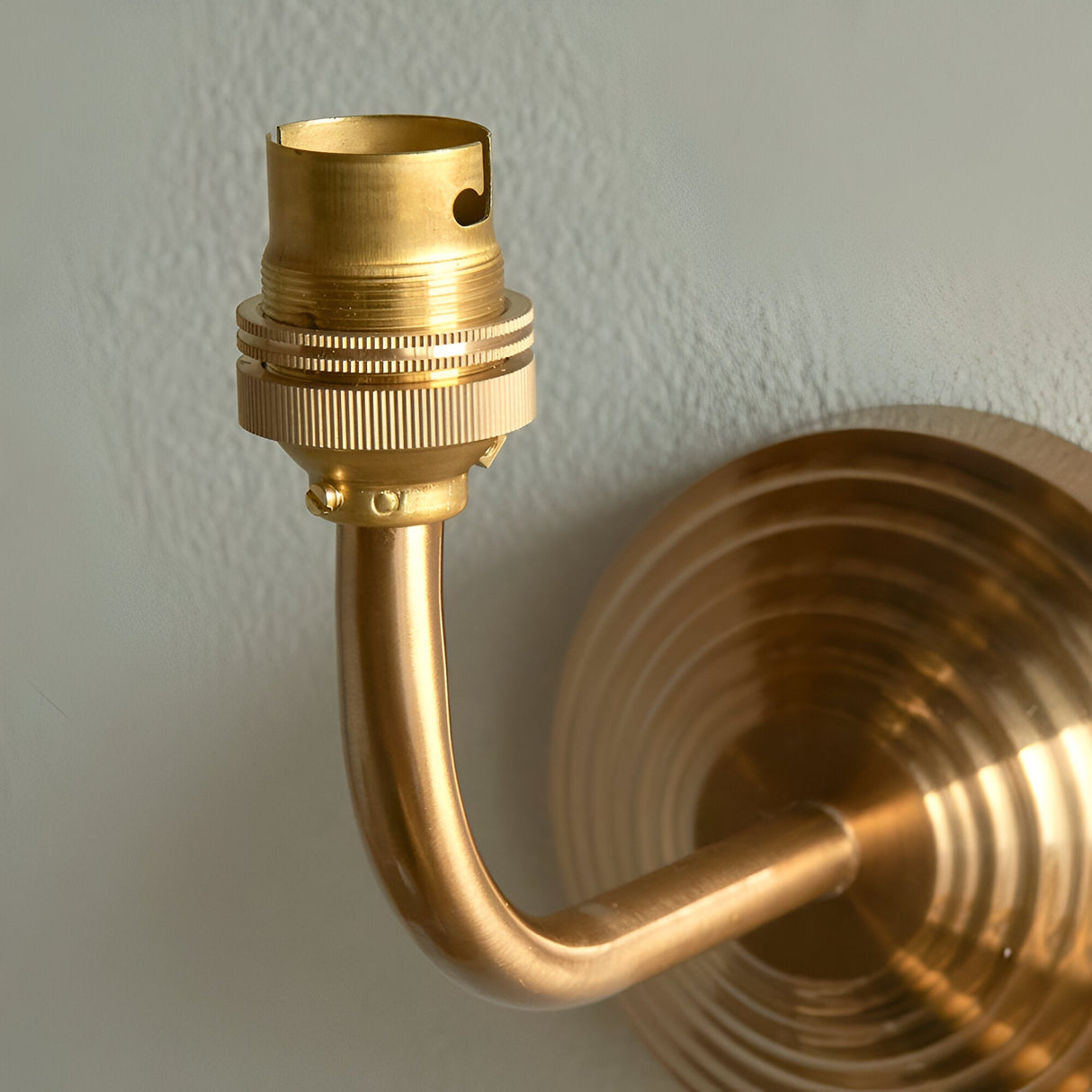 Castgrove wall light in brushed aged brass, close-up of the curved arm and B22 lampholder against a stepped circular wall backplate.