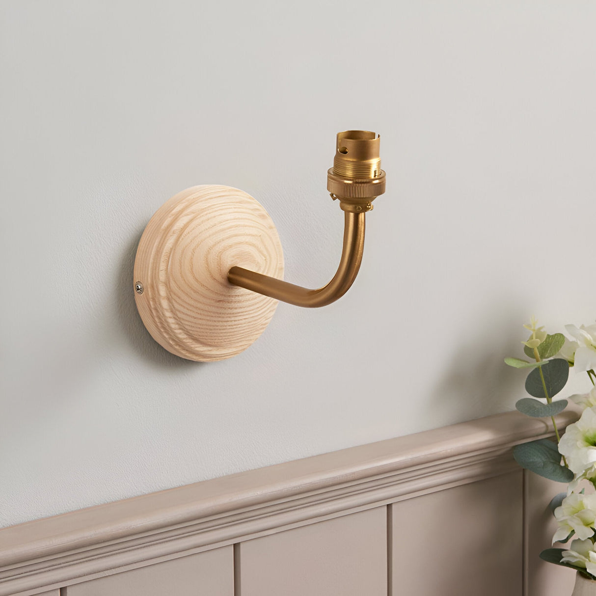 Endon Daneleigh wall light with natural ash wood backplate and curved brushed aged brass arm holding a B22 lampholder, mounted on a light grey wall.