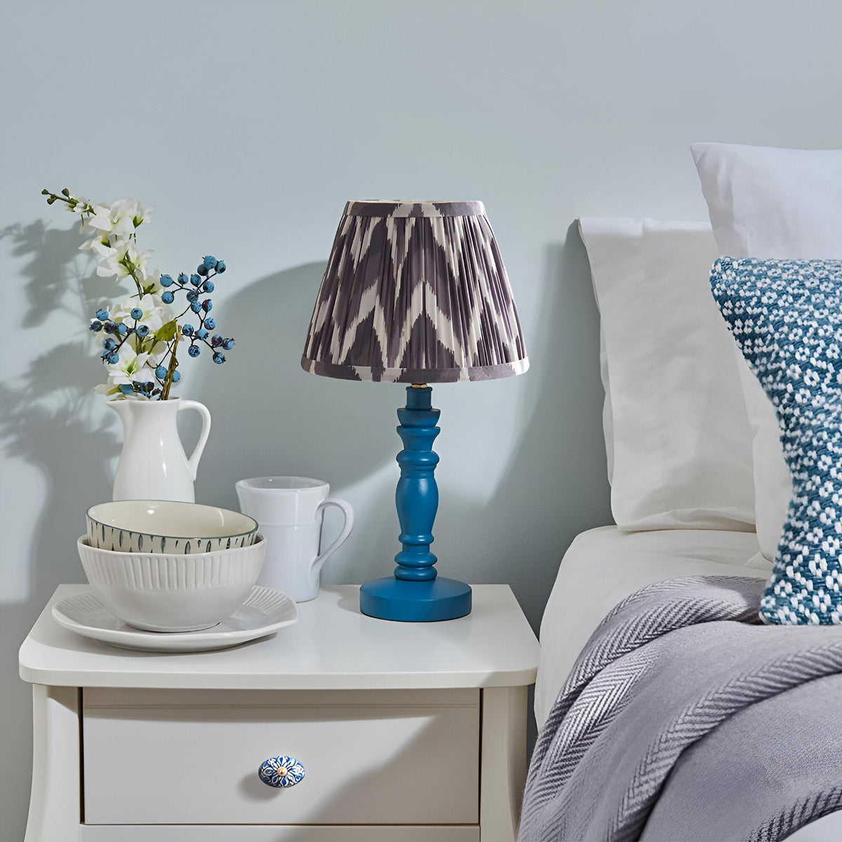 Aldworth Zigzag Table Lamp with turned blue wooden base and grey zigzag fabric shade, styled on a bedside table beside crockery and a bed.