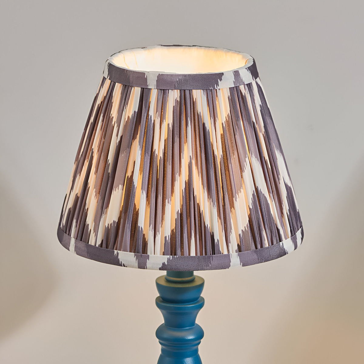 Aldworth Zigzag table lamp with a hand turned wooden spindle base in blue, topped with a pleated grey and cream zigzag print shade, on a plain background.