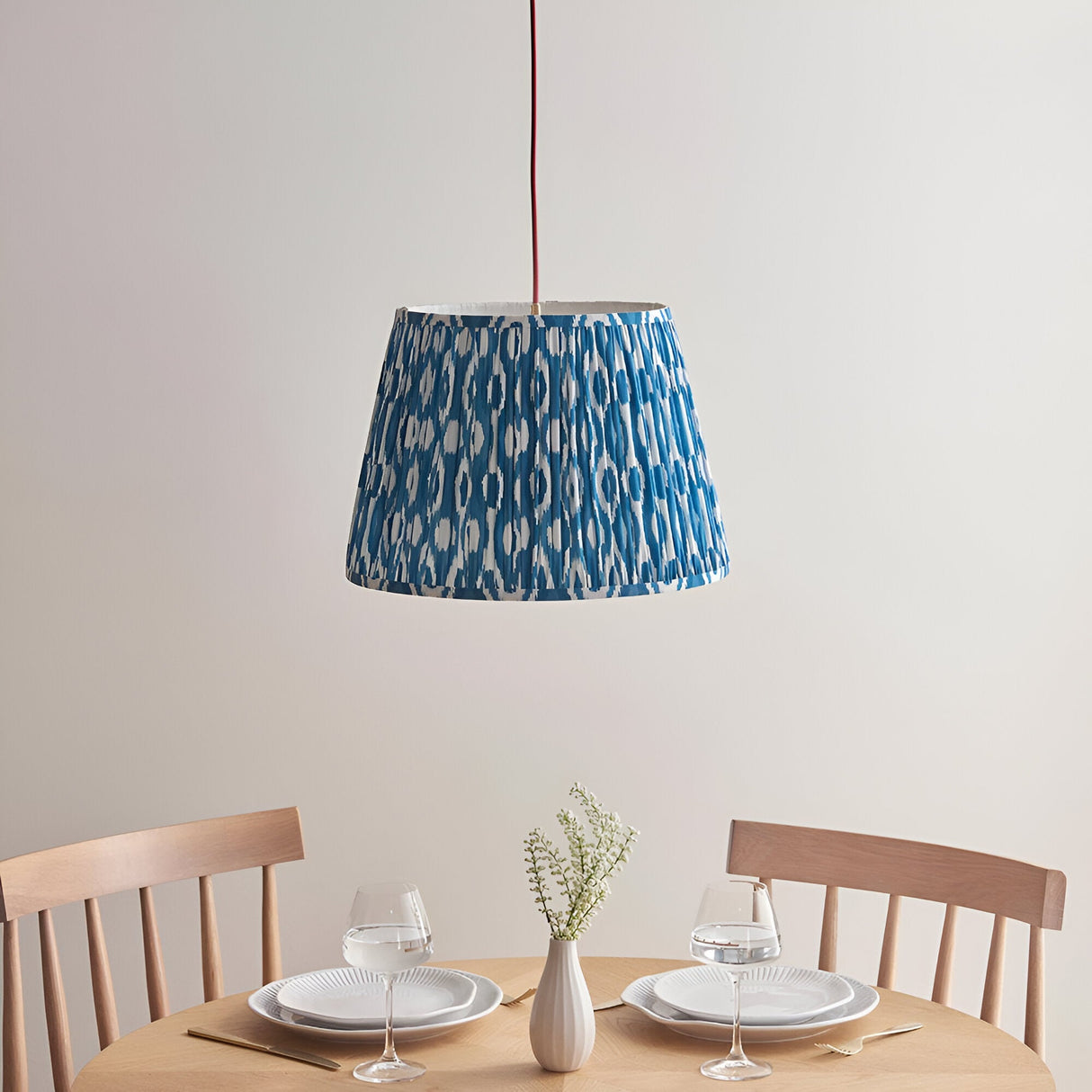 Endon Brackmont Ikat pendant with pleated marlin blue faux silk tapered shade on a red fabric cable, hung above a wooden dining table set for two.