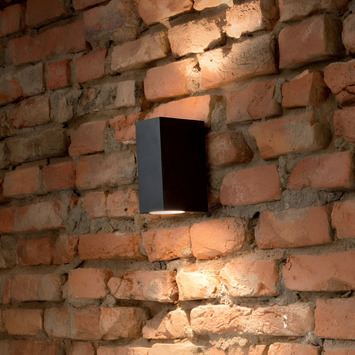 The Aethera Outdoor 2 Light Wall Light - Black casts warm, dimmable lighting onto a rustic red brick wall, accentuating its texture and pattern. Crafted as an outdoor fixture with an IP44 rating for increased durability, it creates a cozy and inviting atmosphere.