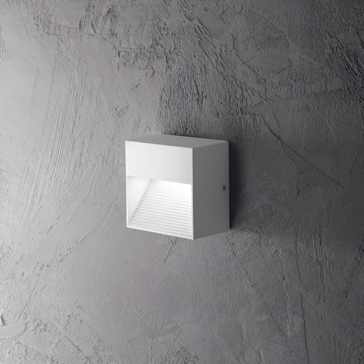 The Jupiter-2 Outdoor Wall Light in white features a modern, minimalist design and is crafted from die-cast aluminum. Mounted on a textured gray wall, its geometric shape includes a horizontal light diffuser that emits a soft, weather-resistant glow downward.