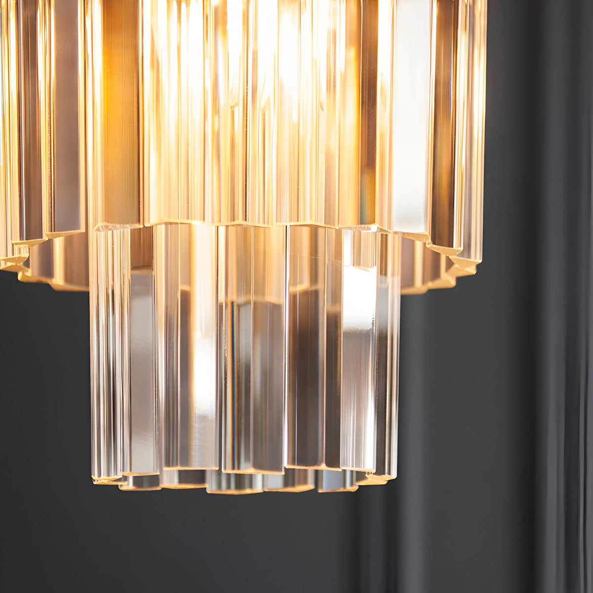 Close-up of Endon Bevstead pendant showing tiered clear lozenge glass drops on a brushed aged brass frame, lit against a dark grey wall.