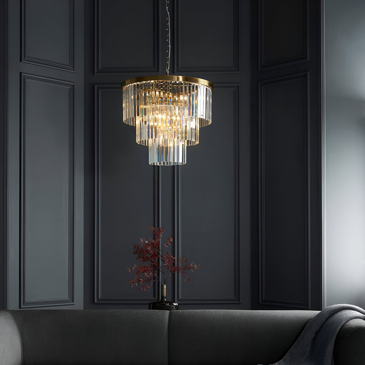Endon Bevstead 7 light pendant in brushed brass with tiered clear crystal drops, hung against a dark panelled wall above a grey sofa.