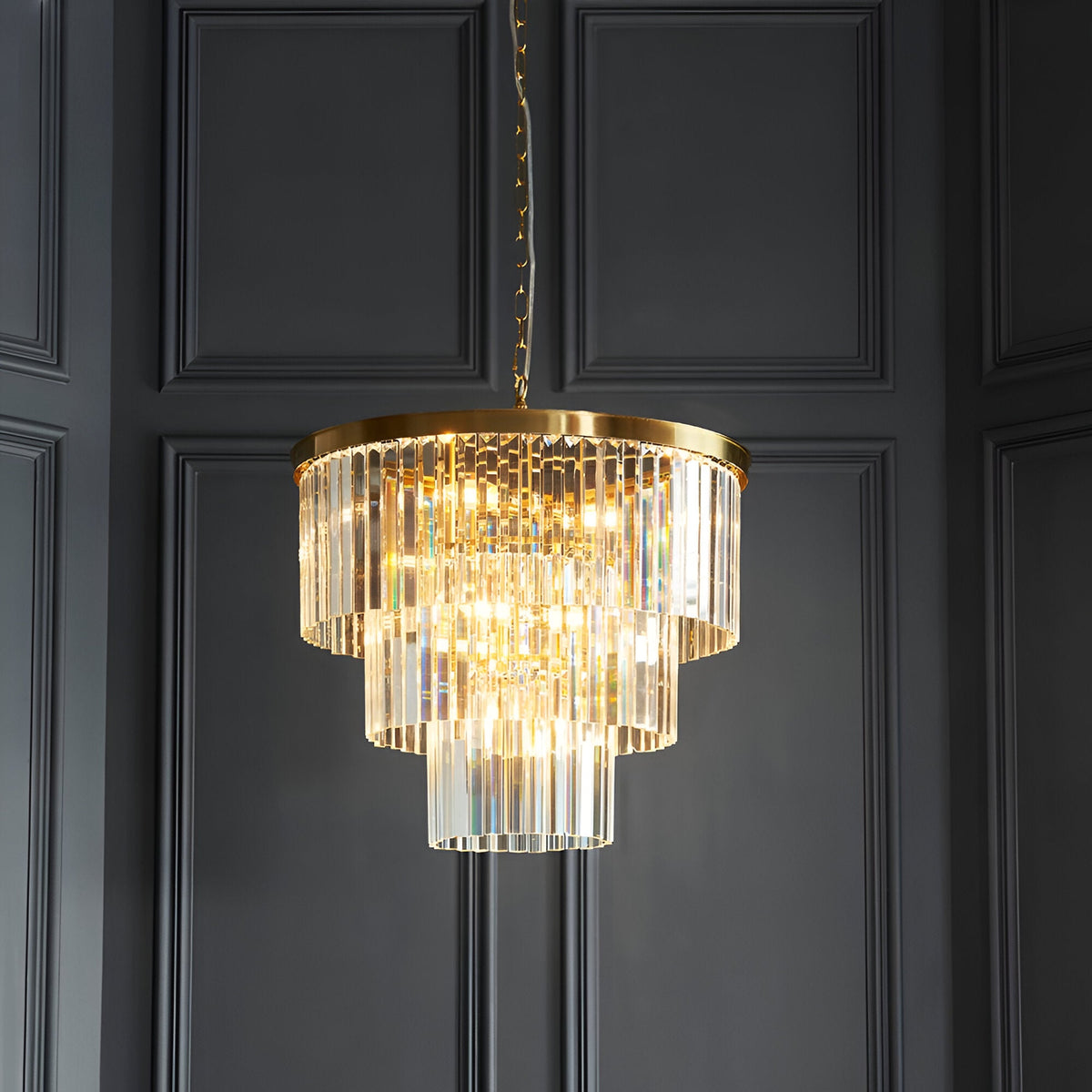 Endon Bevstead 7 light pendant in brushed brass with tiered clear cut glass drops, hung from a chain against dark grey panelled walls.