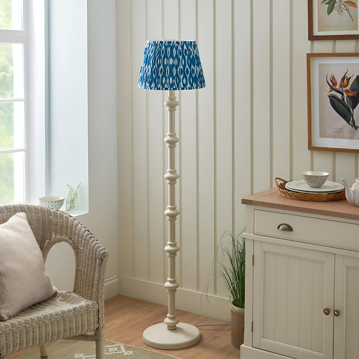 Endon Bevbury Ikat floor lamp with turned white spindle base and blue pleated Ikat patterned shade, styled in a neutral living room beside a wicker chair.