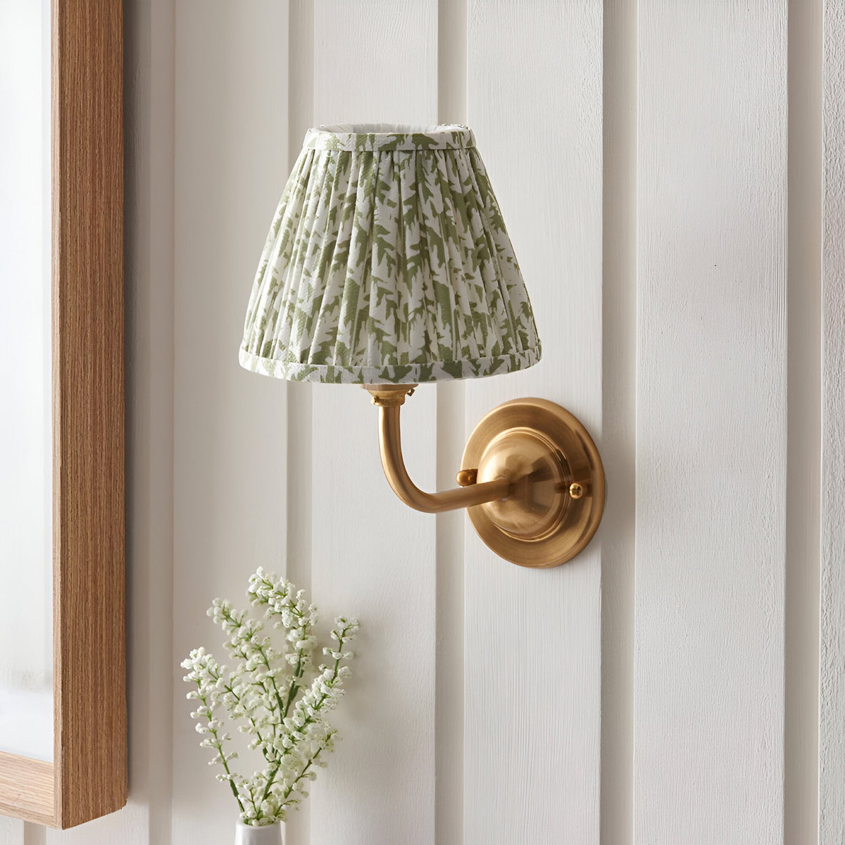 Brackdale Leaf Wall Light with brushed brass curved arm and pleated green leaf-print shade, mounted on a white panelled wall near a mirror and flowers.