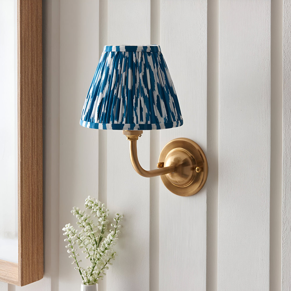 Endon Brackdale Ikat wall light with brushed aged brass curved arm and pleated marlin blue ikat faux silk shade, mounted on a white panelled wall.