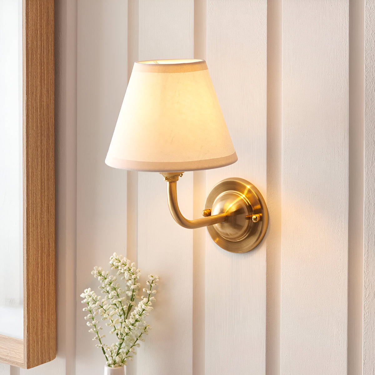 Brackdale Ivy Wall Light in Vintage White with a tapered fabric shade and curved brushed brass arm, mounted on a panelled wall above white flowers.