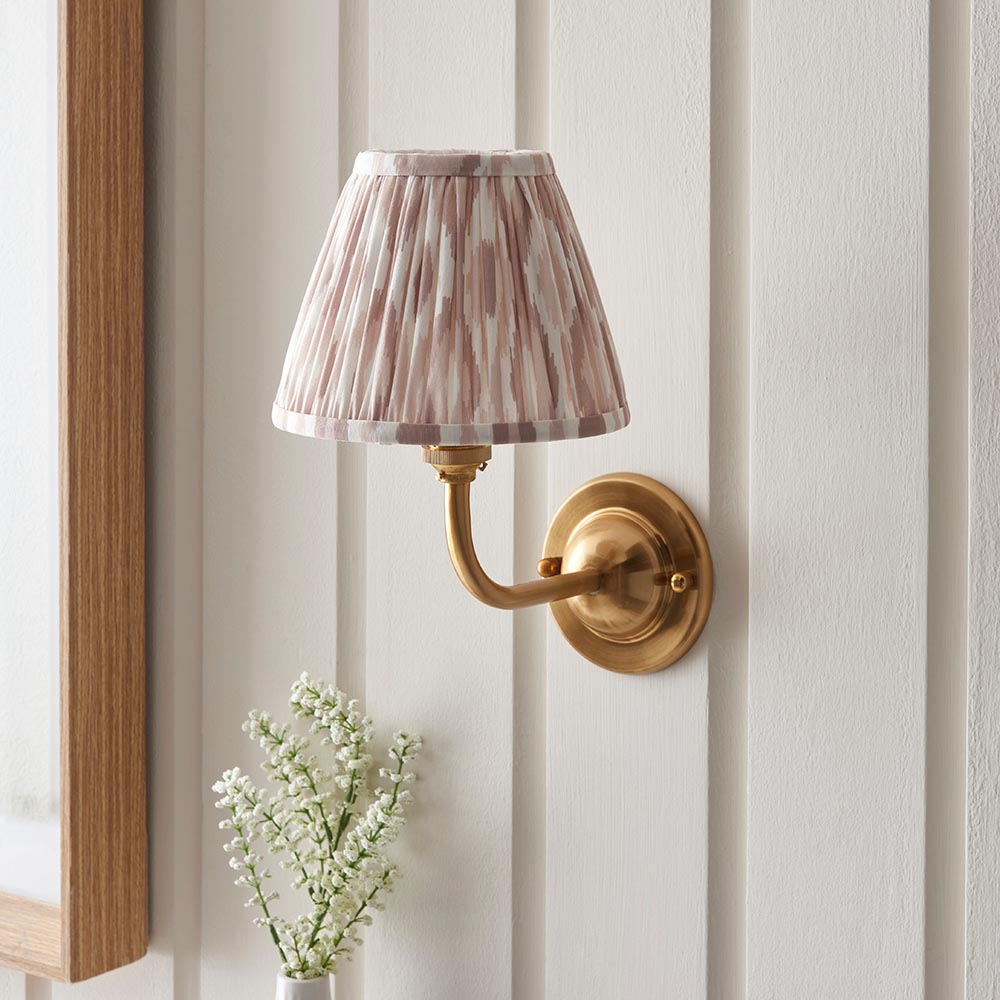 Endon Brackdale Ikat wall light with a brushed aged brass curved arm and dome, topped with a pleated pink and natural Ikat shade, mounted on panelled wall.