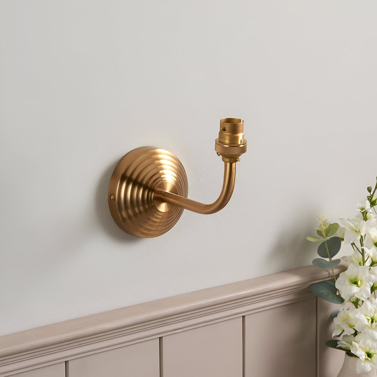 Endon Castgrove Ripple wall light with stepped brushed aged brass backplate, curved arm and B22 lamp holder, mounted on a pale wall above panelling.