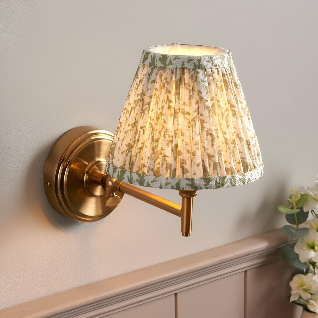 Cliffwood Leaf Wall Light with brushed brass knuckle arm and pleated fabric shade in cream with green leaf print, mounted on a light grey wall.