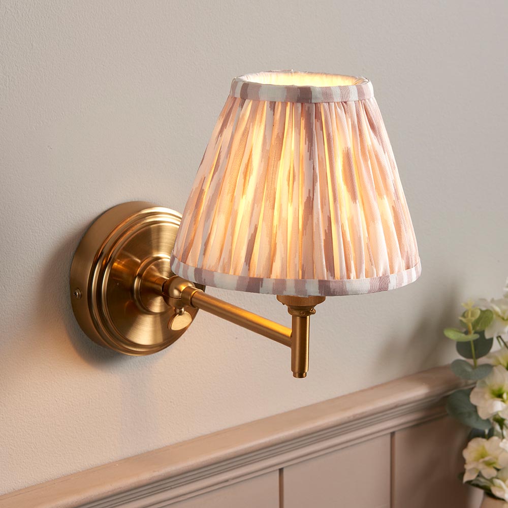 Cliffwood Ikat Wall Light in brushed brass with an adjustable arm and pleated natural pink-toned Ikat fabric shade, mounted on a light grey wall.