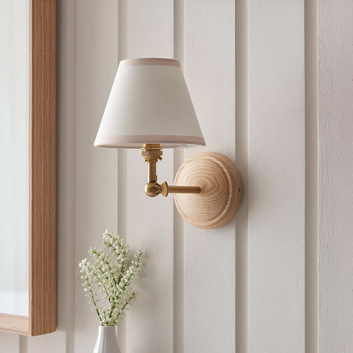 Danemere Ivy wall light with tapered fabric shade, brass fittings and ash wood backplate, shown mounted on a panelled wall beside a mirror and vase of flowers.