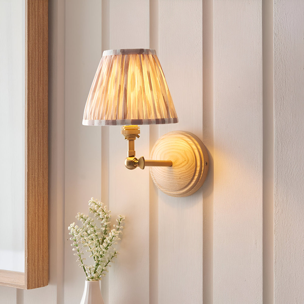 Endon Danemere wall light with natural ash wood back plate, brushed aged brass arm and pleated Ikat faux silk shade, mounted on cream panelling.