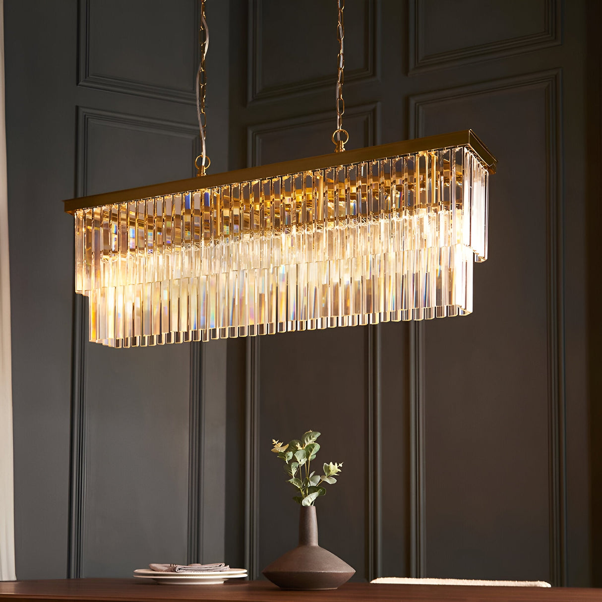 Bevstead 4-light linear pendant with brass band and tiered crystal glass drops, hung on twin chains above a dining table in a dark-panelled room.