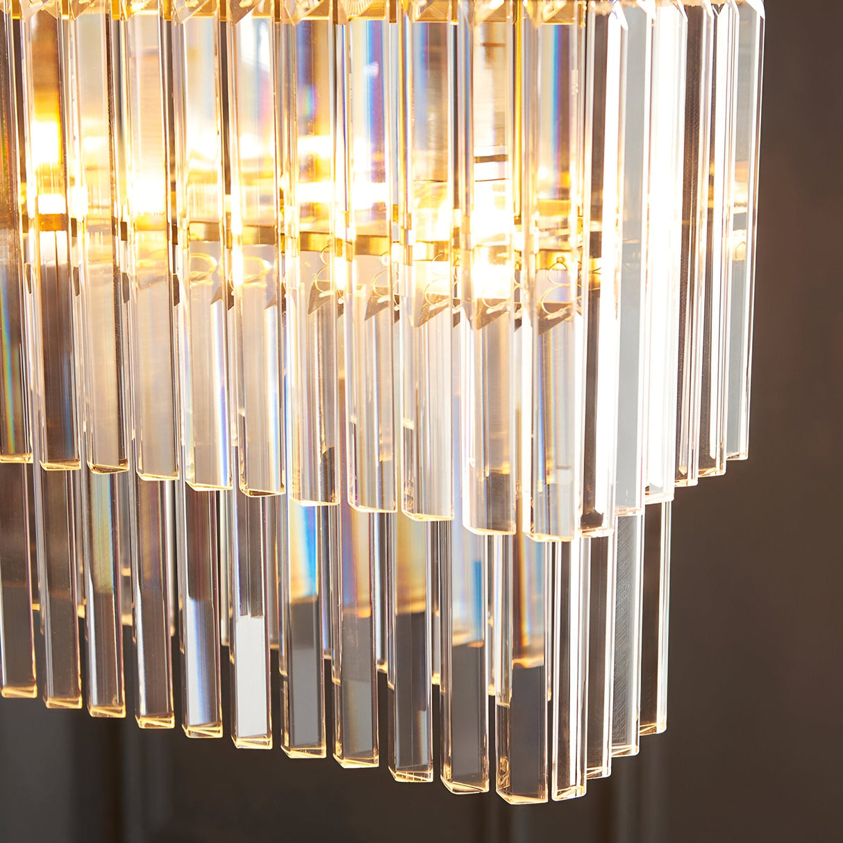 Close-up of the Bevstead 4 Light Linear Pendant in brushed brass, showing tiered clear lozenge glass drops illuminated by warm bulbs.