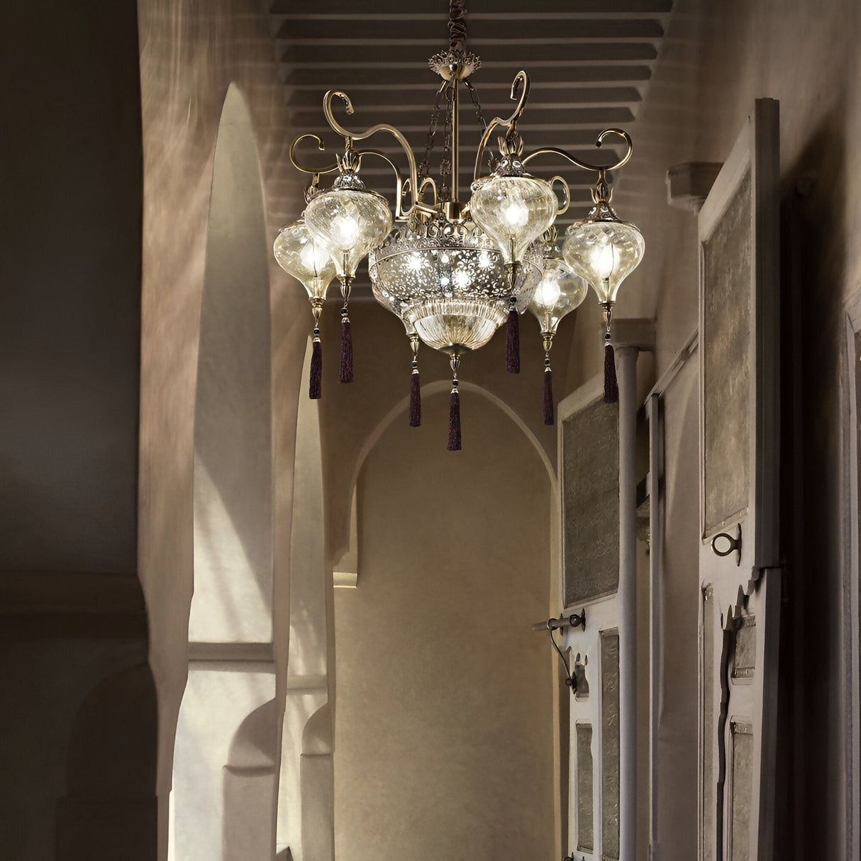 The Daley Crystal 9 Light Chandelier - Brass, with glass globes and crystal accents, hangs in a dim hallway featuring arched doorways and rustic walls. Its soft glow casts warm shadows, creating an inviting, elegant ambiance.