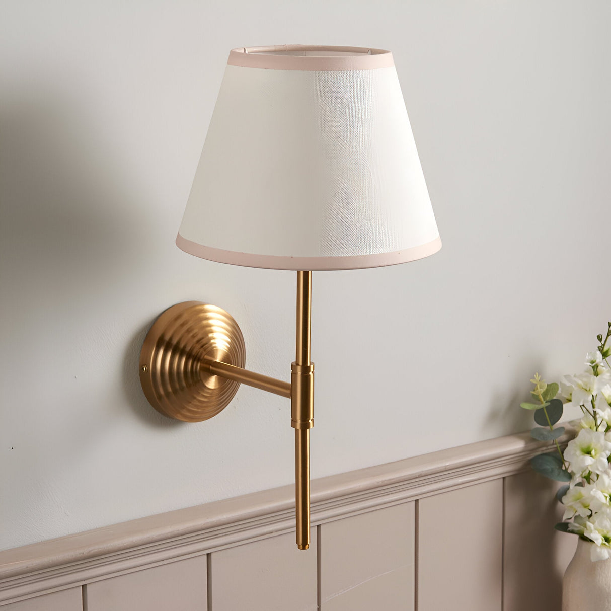 Castleigh Ivy wall light with antique brass T-shaped arm, ribbed circular backplate, and tapered white shade with pink trim, shown mounted on a light wall.