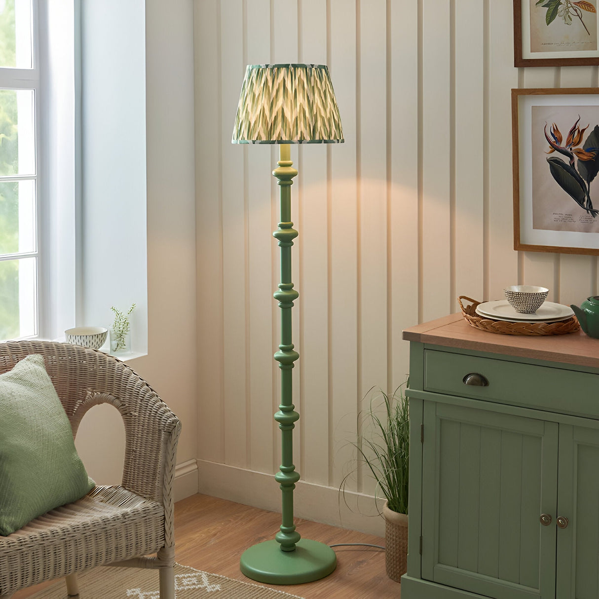 Bevbury Zigzag Floor Lamp with turned wooden green base and pleated zigzag-patterned shade, shown in a living room beside a wicker chair and green sideboard.