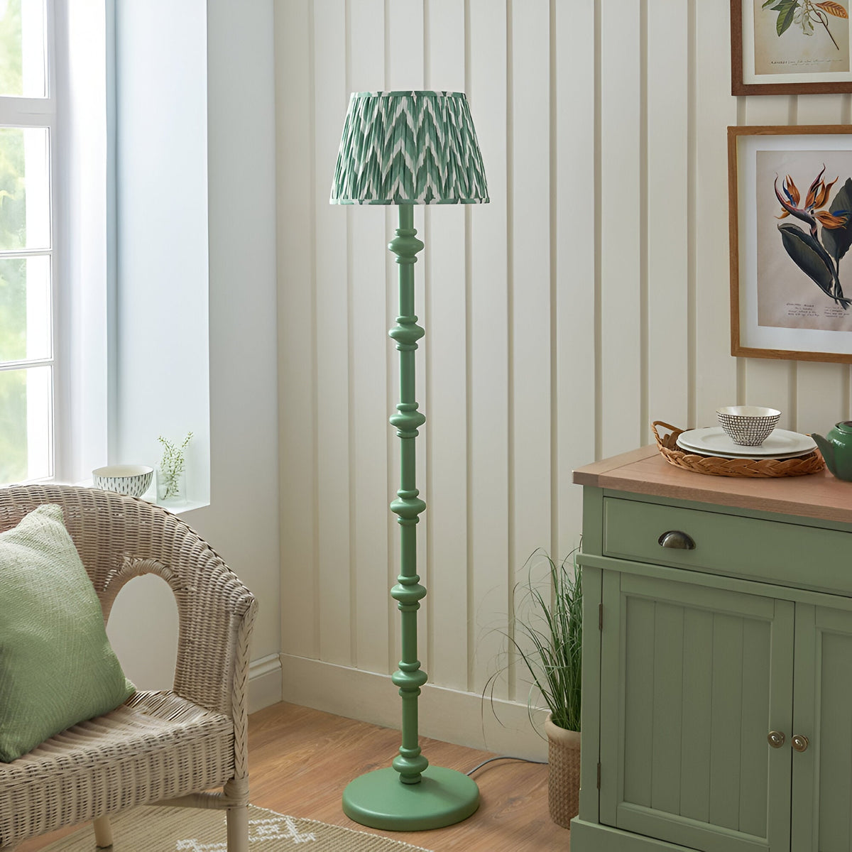 Bevbury Zigzag Floor Lamp with green turned wooden spindle base and green zigzag patterned shade, shown beside a wicker chair in a living room.