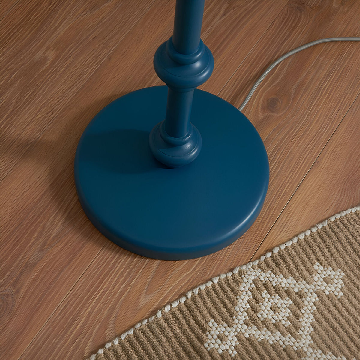 Close-up of the Endon Bevbury Ripple floor lamp's marlin blue turned wooden spindle base with grey braided flex on wood flooring beside a woven rug.