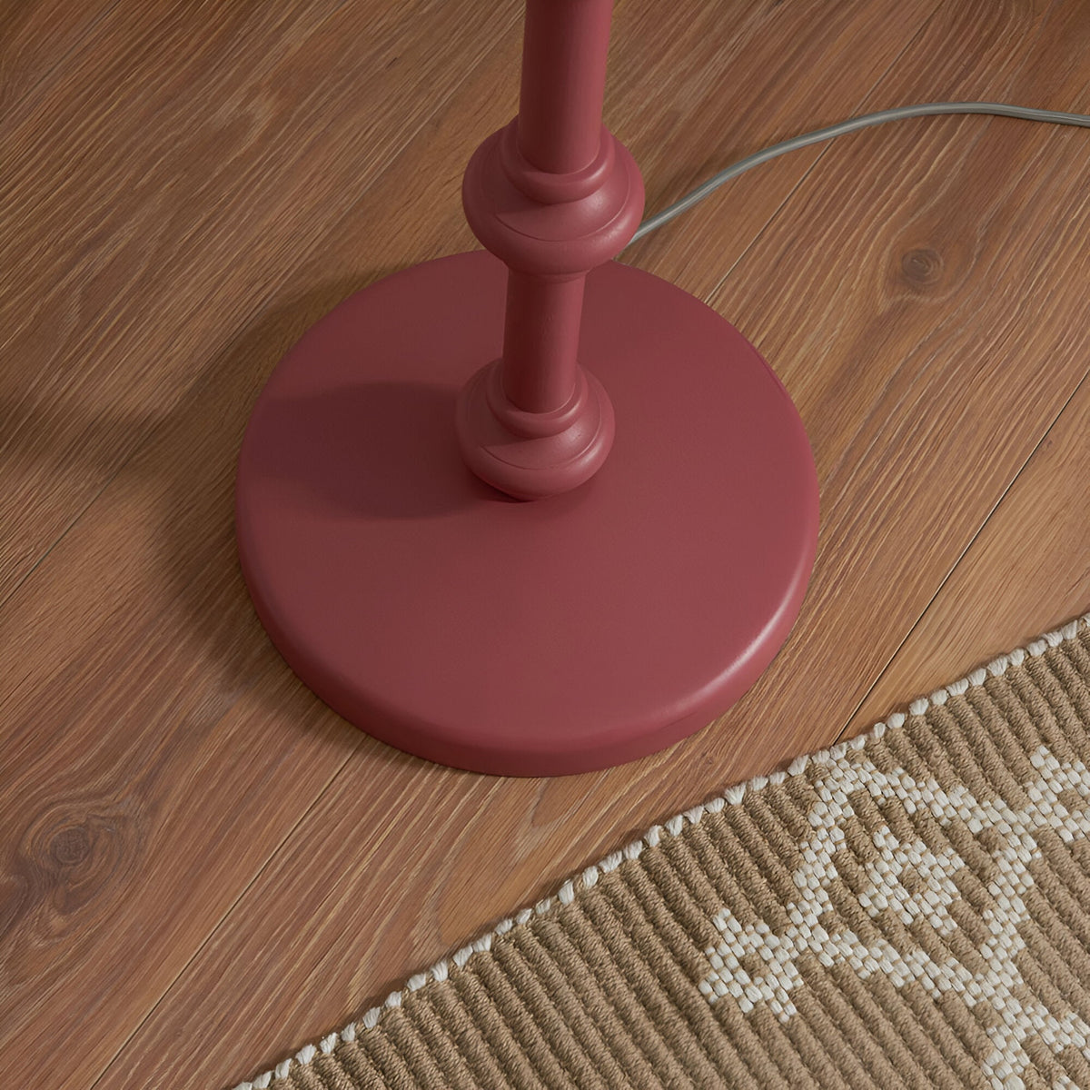 Close-up of the Endon Bevbury floor lamp's pink turned wood spindle base on wooden flooring beside a woven rug, with grey braided flex trailing.
