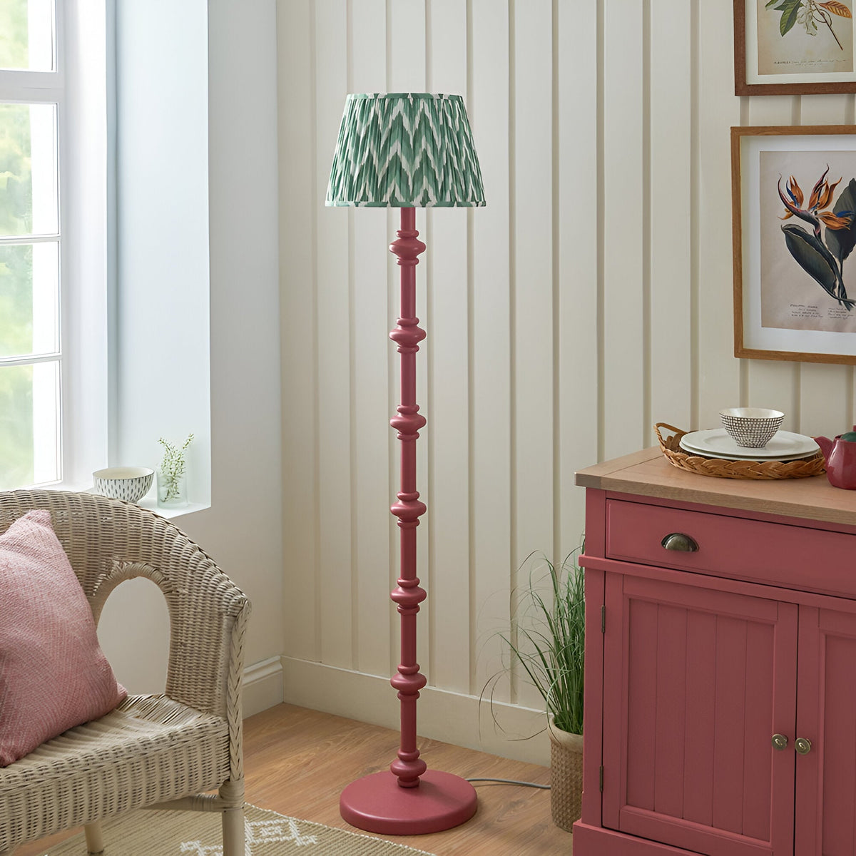 Endon Bevbury floor lamp with pink clay turned wood spindle base and green zigzag pleated shade, styled beside a wicker chair and pink sideboard.