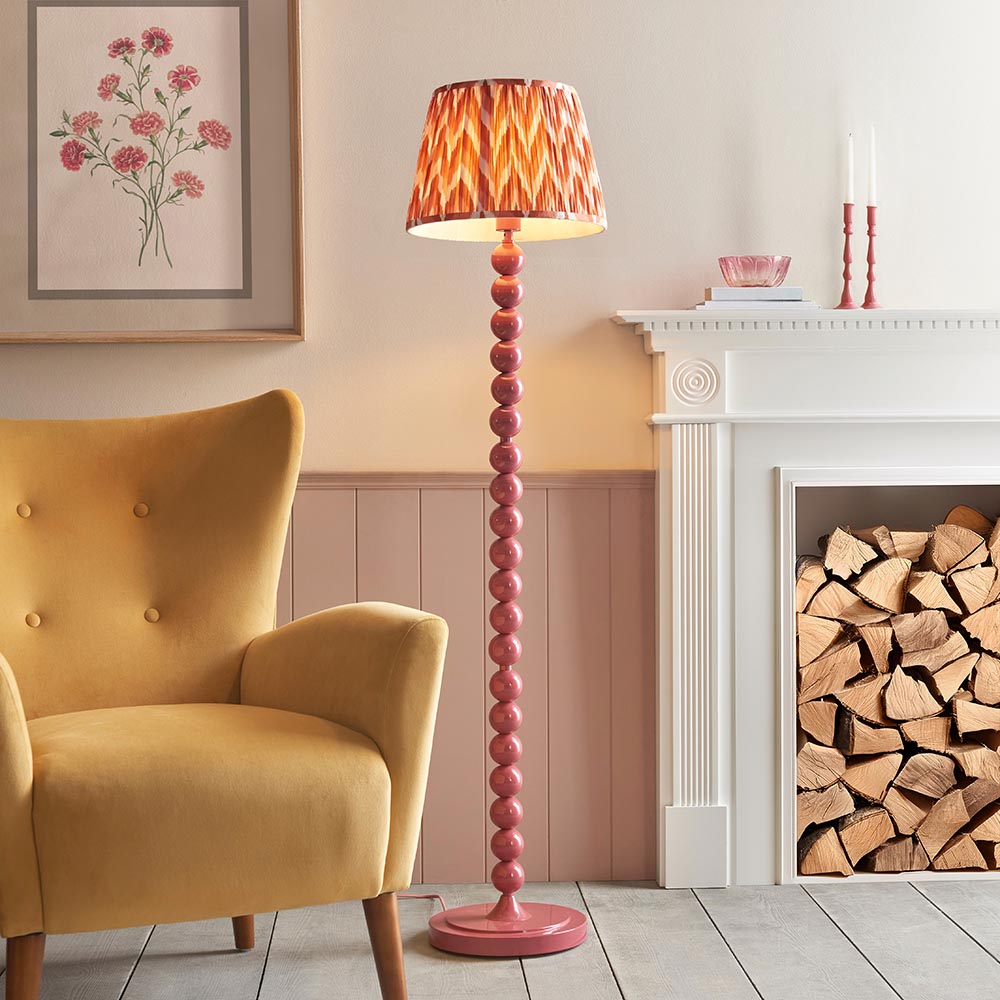 Brenleigh Zigzag Floor Lamp in pink with stacked sphere metal base and pleated orange zigzag fabric shade, shown beside a mustard armchair and fireplace.