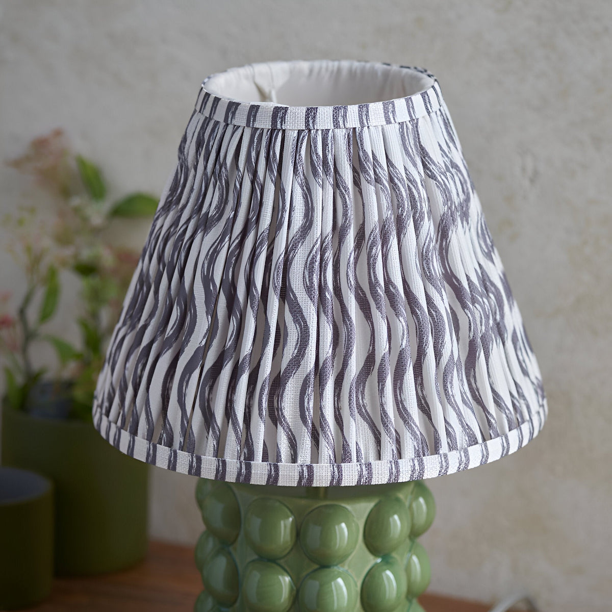 Endon Amberley Ripple table lamp with olive green ceramic bobble base and pleated grey wave-pattern fabric shade, shown on a wooden surface.