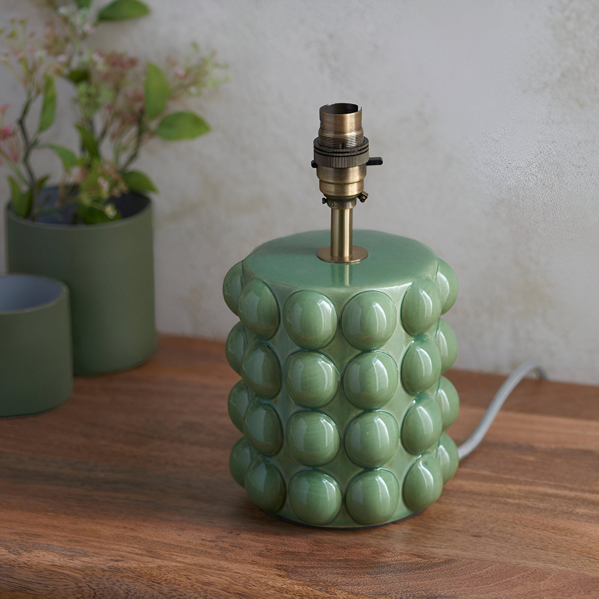 Endon Amberley Ripple table lamp base in gloss olive green ceramic with bobble texture and aged brass B22 holder, on a wooden surface beside plant pots.
