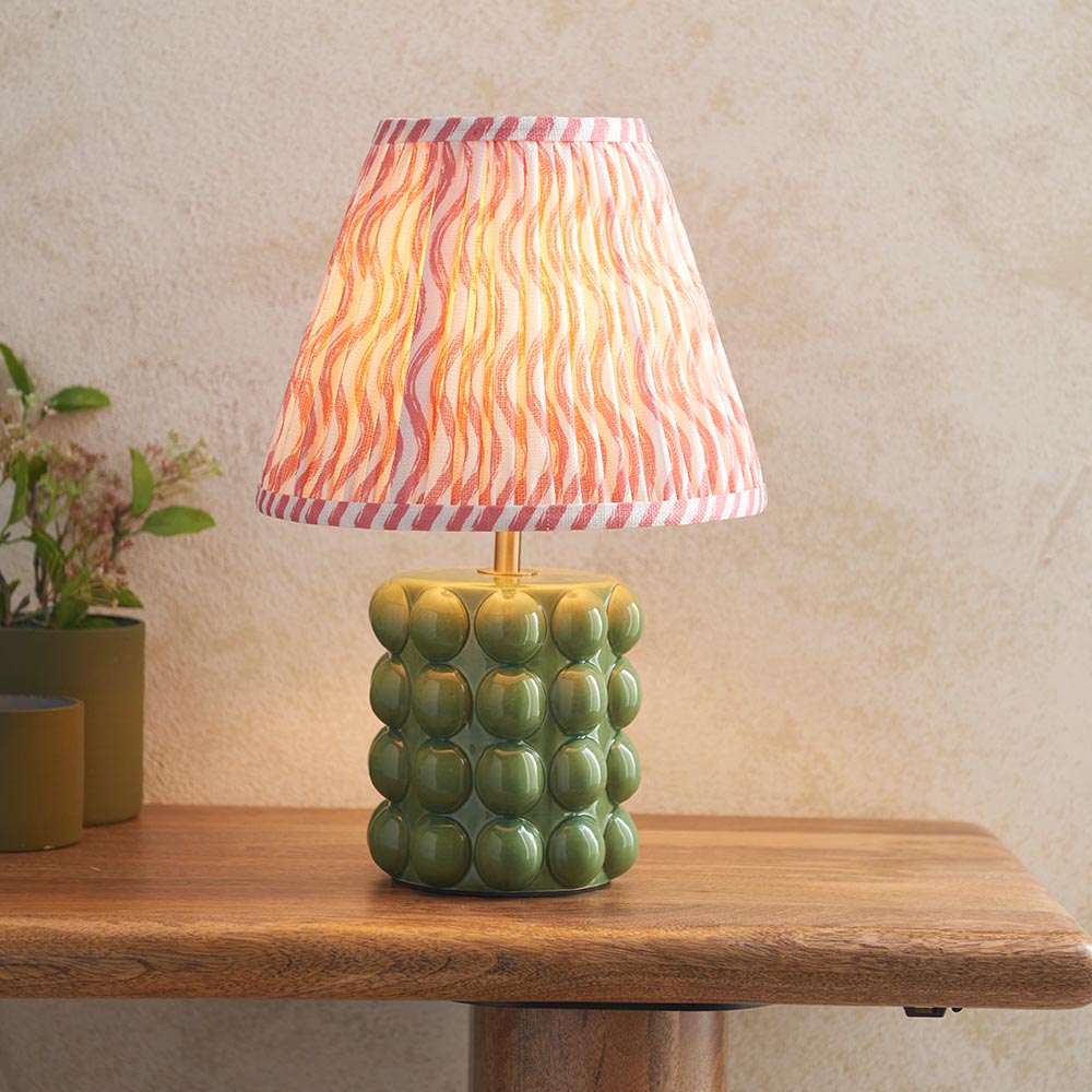 Amberley Ripple Table Lamp with green ceramic bobble base and pink wavy-print fabric shade, shown lit on a wooden table beside potted plants.
