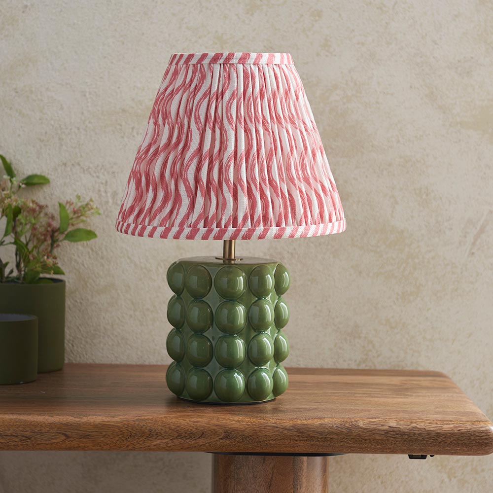 Amberley Ripple Table Lamp with a green ceramic bobble base and pink and white wavy-striped pleated shade, shown on a wooden table beside potted plants.