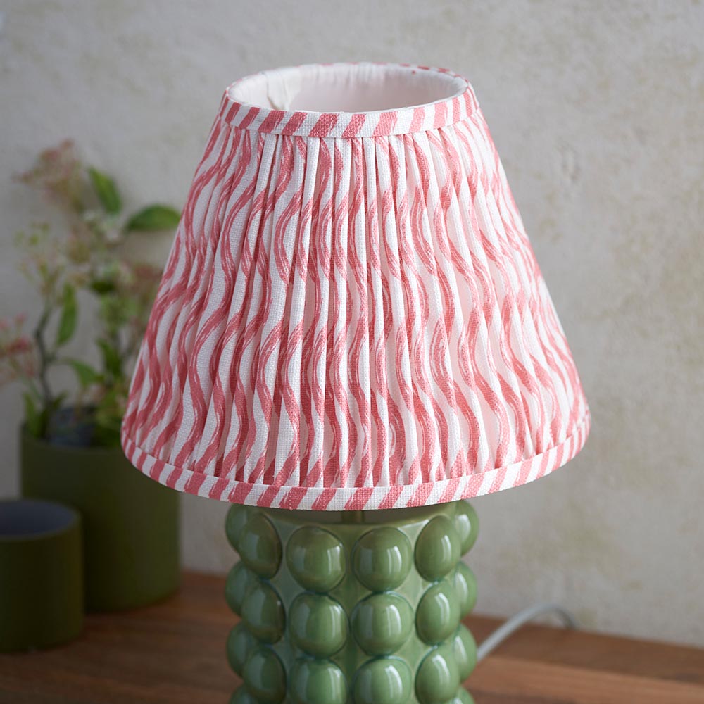 Amberley Ripple Table Lamp with green ceramic bobble base and pink-and-white ripple pattern pleated shade, shown beside potted plants on a wooden surface.