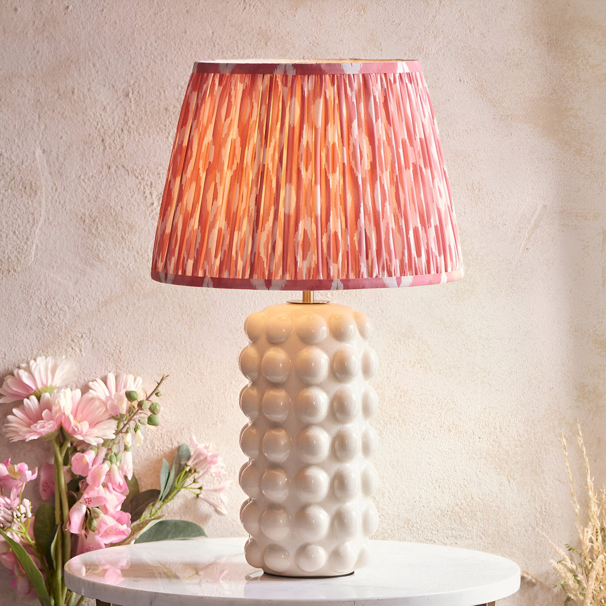 Amberley Ikat table lamp with a bobble-textured white ceramic base and pleated pink, orange and grey Ikat-print shade on a marble table.
