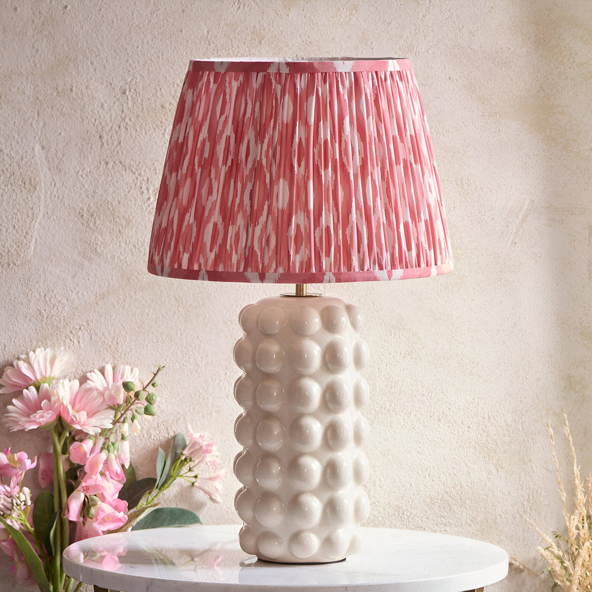 Amberley Ikat Table Lamp with white ceramic bobble base and pink Ikat patterned shade, shown on a marble side table beside pink flowers.