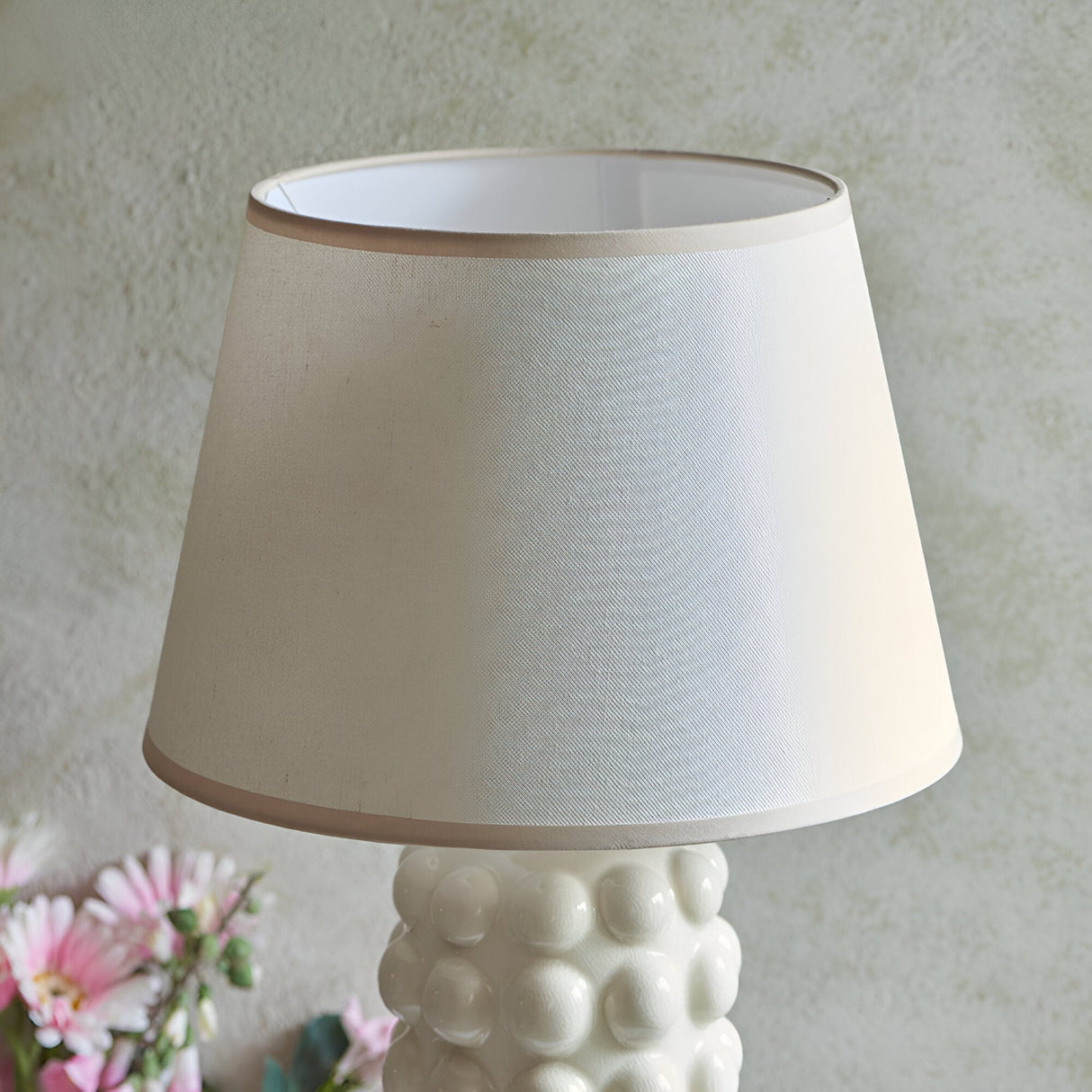Endon Amberley Ivy table lamp shade in vintage white cotton-mix fabric with taped edge, shown above the bobble ceramic base beside pink flowers.