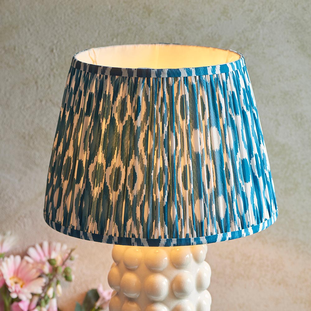 Endon Amberley Ikat table lamp lit on a white ceramic bobble base, with a pleated blue, grey and white ikat shade beside pink flowers.