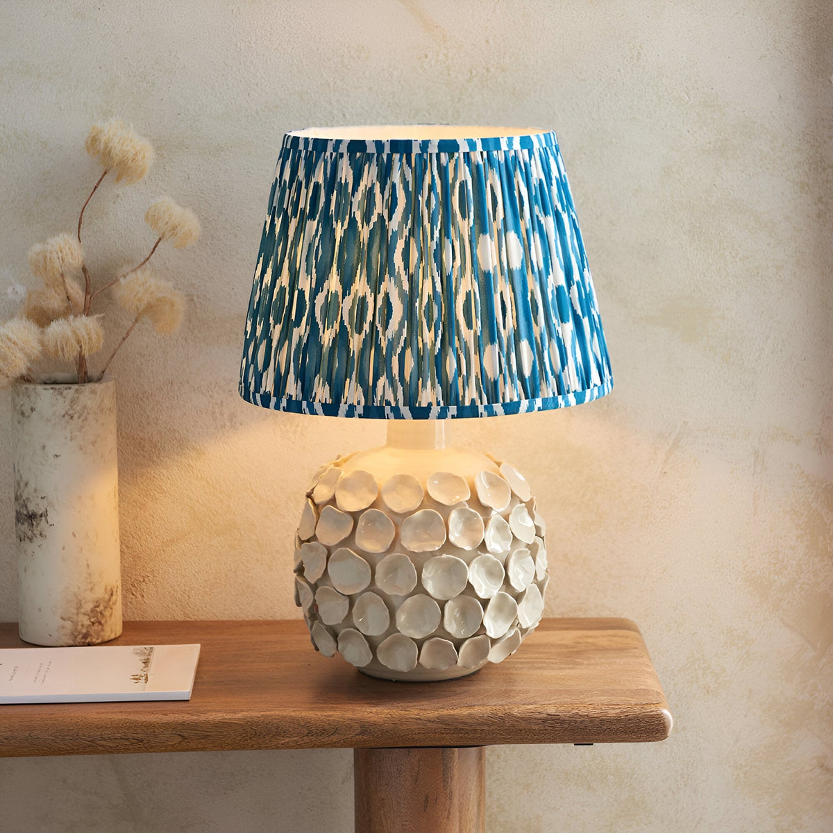 Ashbury Ikat Table Lamp with ivory crackle-glaze ceramic base and pleated blue patterned shade, shown lit on a wooden table beside dried flowers.