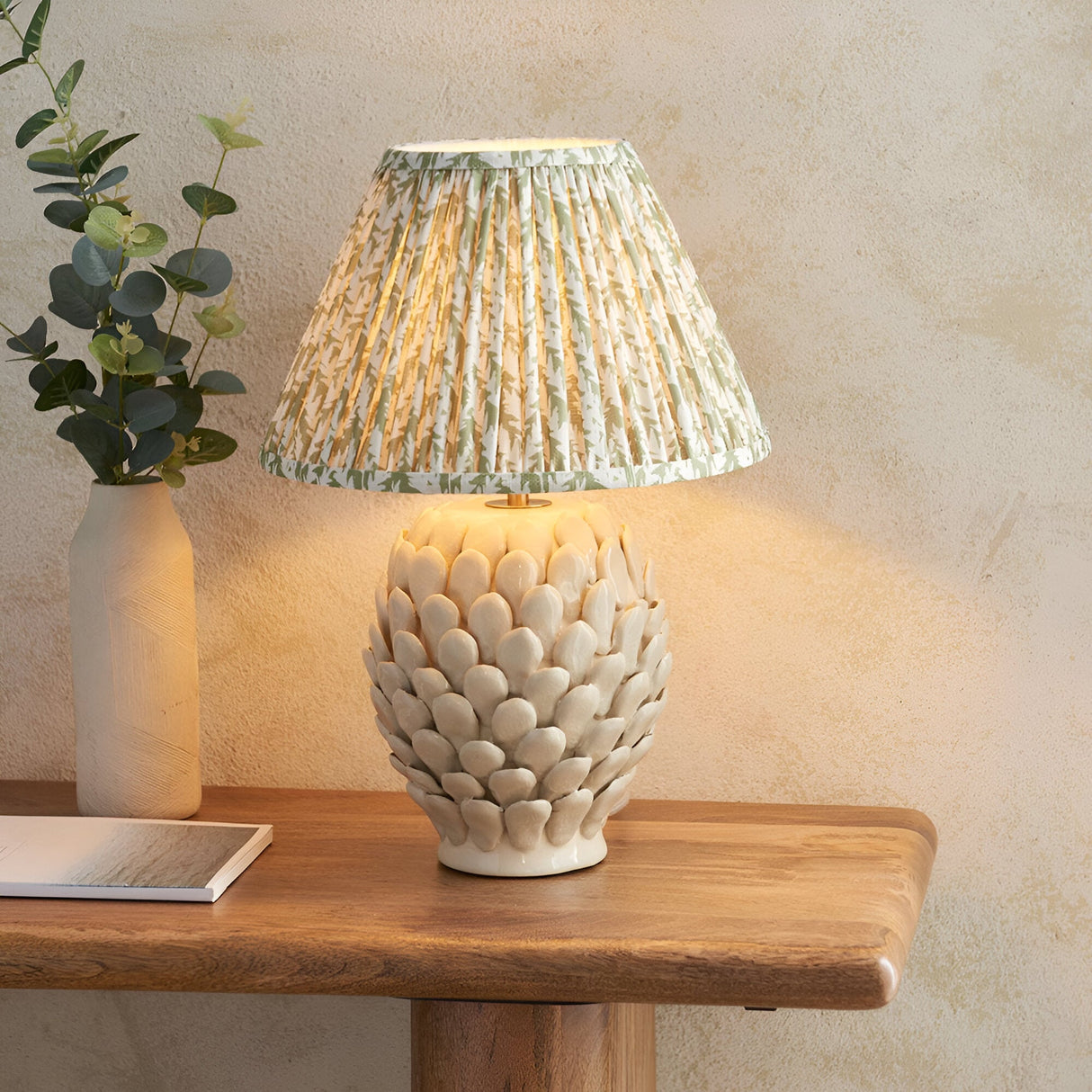 Endon Caldford table lamp with ivory crackle-glazed ceramic artichoke base and pleated green leaf-print shade on a wooden console beside a eucalyptus vase.