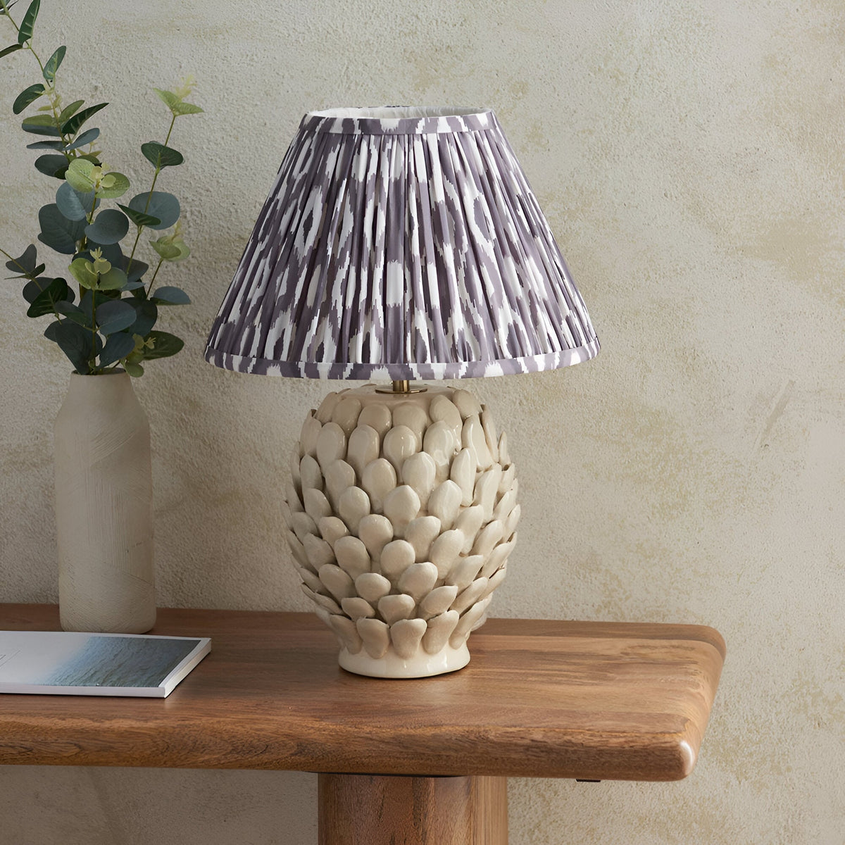 Endon Caldford table lamp with ivory ceramic leaf-textured base and pleated grey Ikat shade, styled on a wood console with a eucalyptus vase.