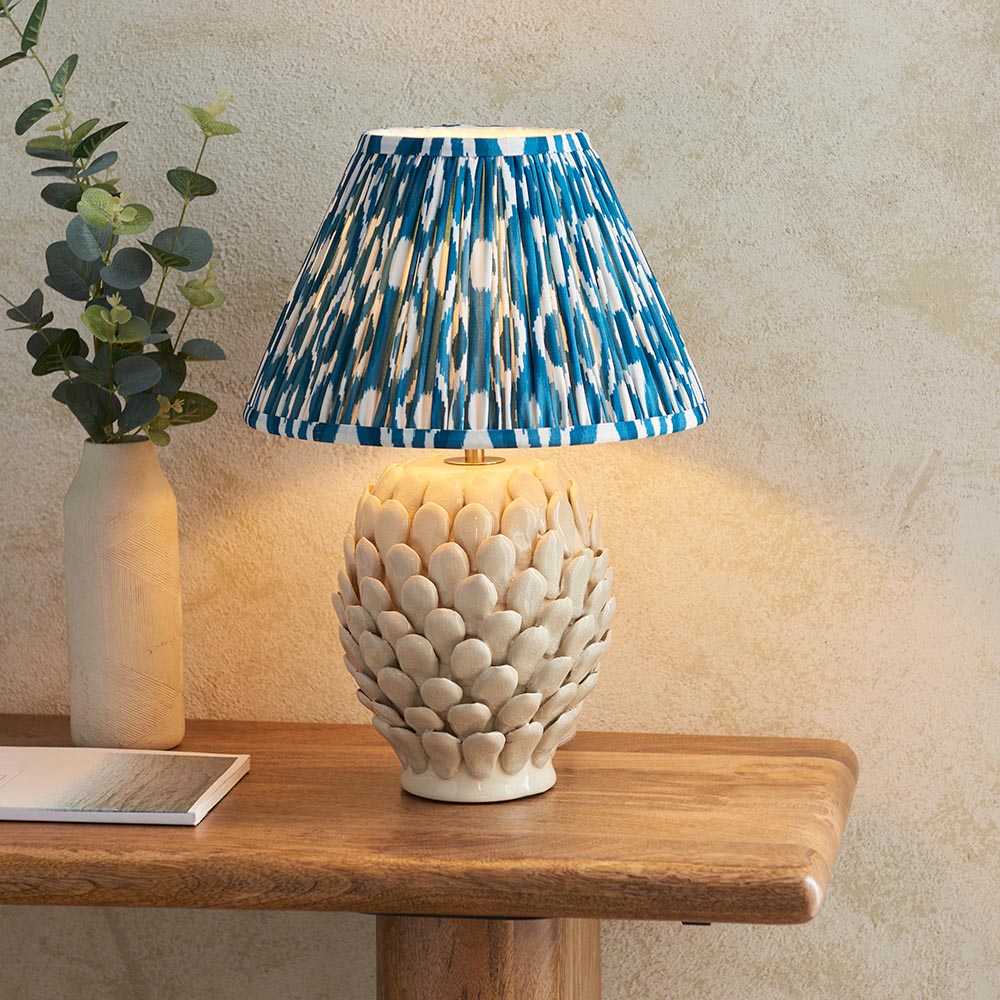Caldford Ikat 30cm Table Lamp with cream ceramic leaf-detail base and blue patterned fabric shade, shown lit on a wooden table beside a vase.