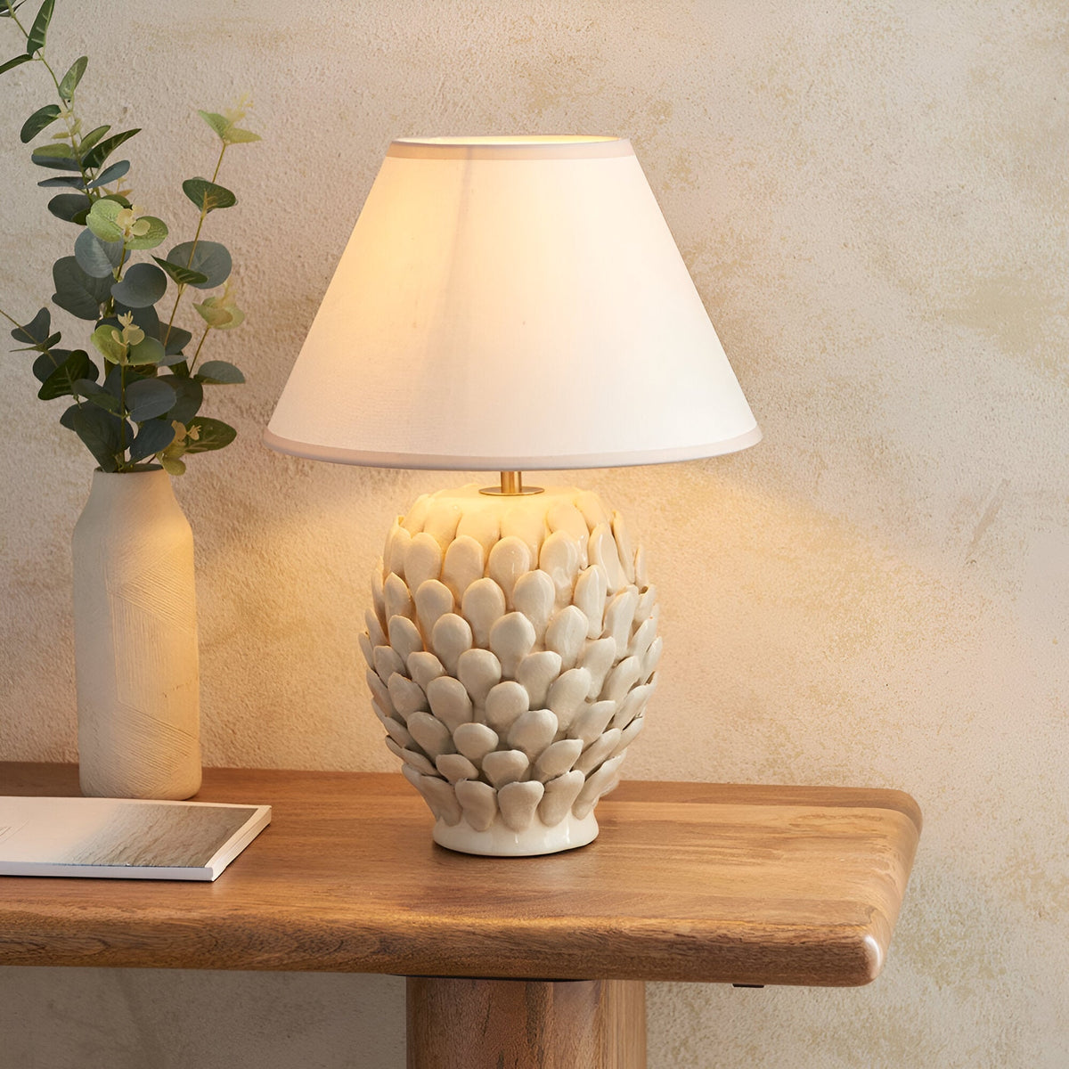 Caldford Ivy 30cm table lamp with white leaf-textured ceramic base and white tapered fabric shade, shown lit on a wooden console table.