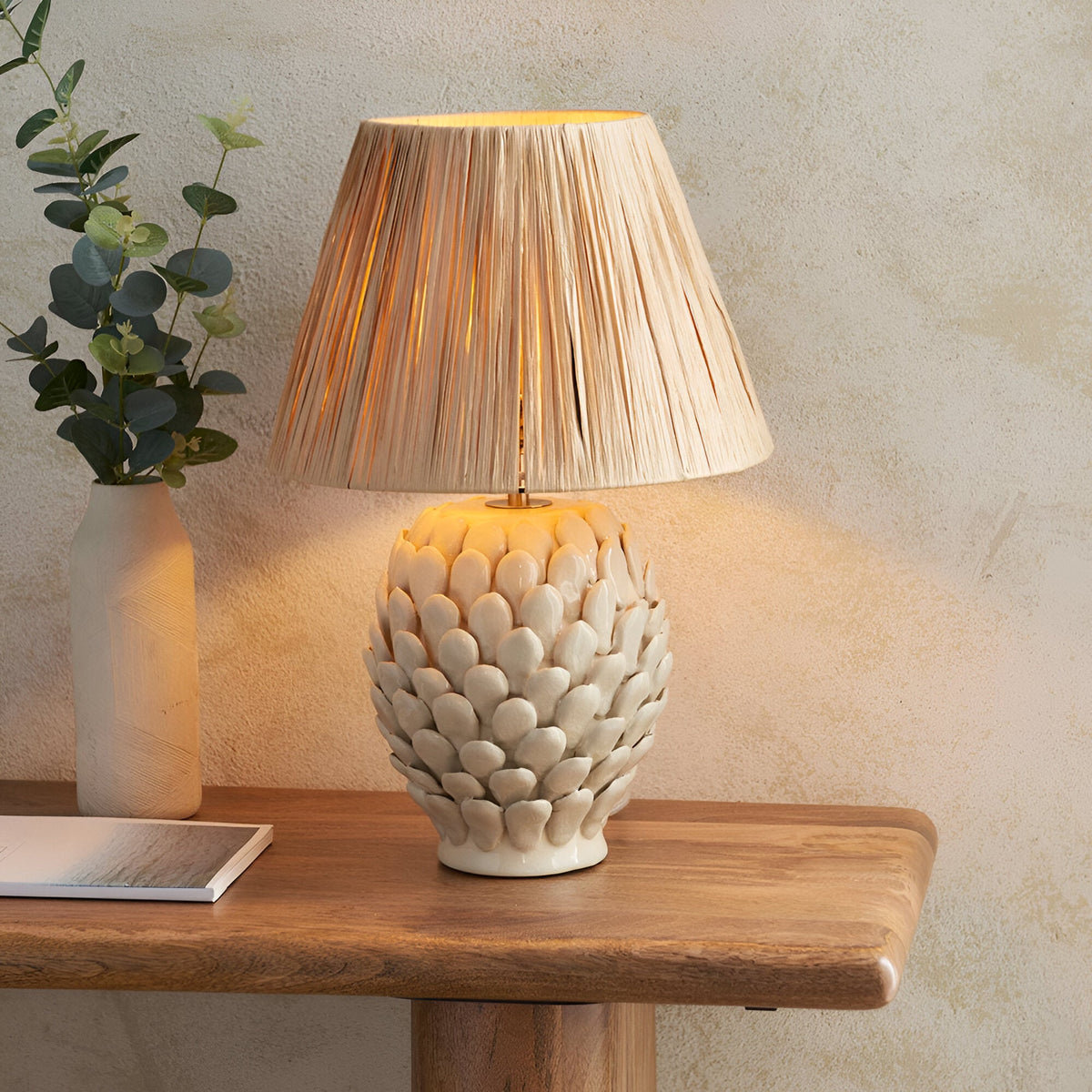 Endon Caldford table lamp with ivory crackle-glazed ceramic artichoke base and natural raffia tapered shade, lit on a wooden console beside a eucalyptus vase.