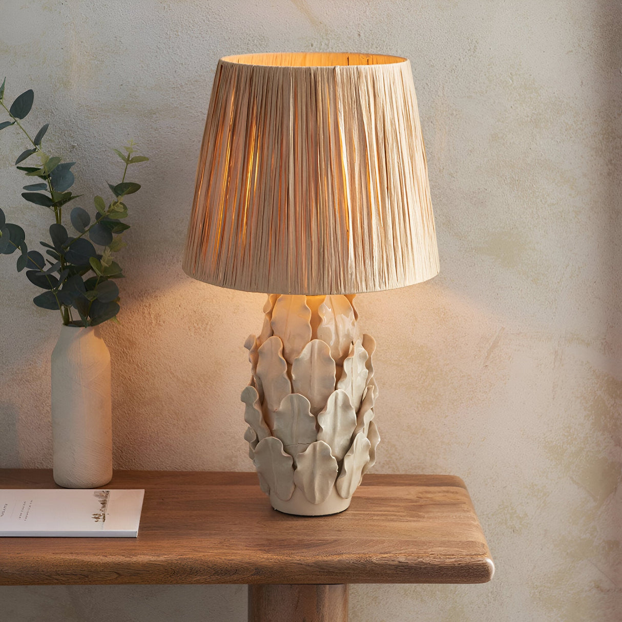 Endon Caldford table lamp with ivory crackle-glazed ceramic leaf base and natural raffia tapered shade, lit on a wood console beside a eucalyptus vase.