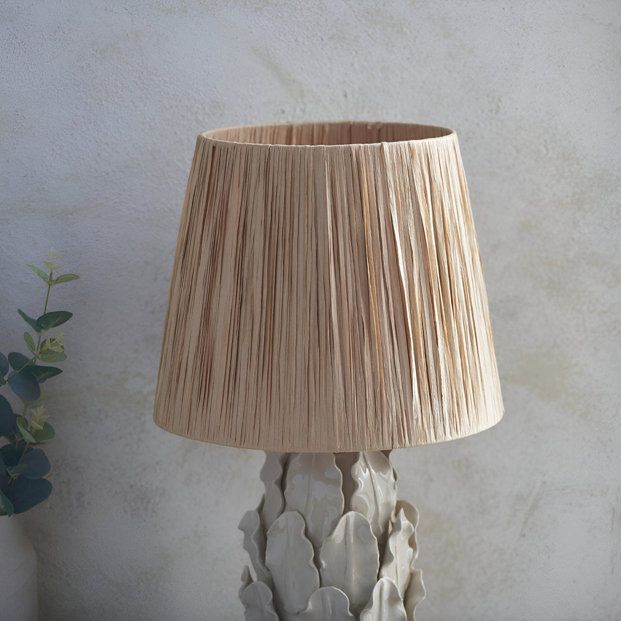 Endon Caldford table lamp with tapered natural raffia shade and ivory crackle-glazed ceramic leaf base, beside eucalyptus against a pale textured wall.